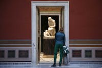 Una visitatrice al Teatro Torlonia di Roma che indossa una camicia blu e pantaloni verde. La stanza ha le pareti bordeaux con una porta di marmo aperta che lascia intravedere una scultura