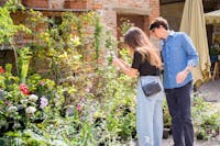 Una coppia di visitatori che stanno fotografando alcuni fiori di uno stand di Herbarium. La ragazza ha una maglietta e borsa nera e un paio di jeans chiari, mentre il ragazzo ha capelli corti neri, occhiali da sole, jeans scuri e camicia di jeans